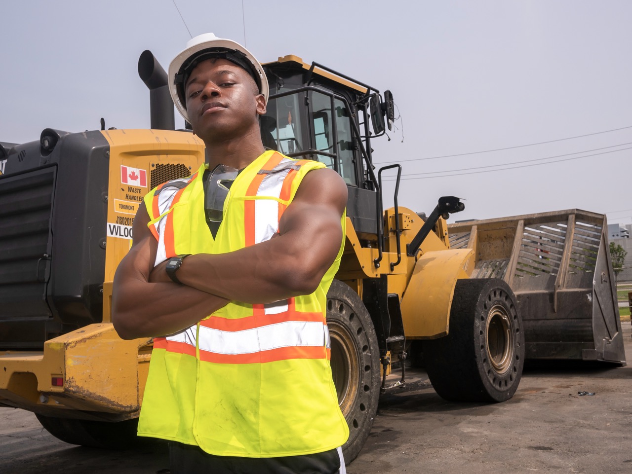 Confident construction worker in helmet and yellow safety vest stands with arms crossed near yellow heavy machinery. GFL Property Toronto, Ontario